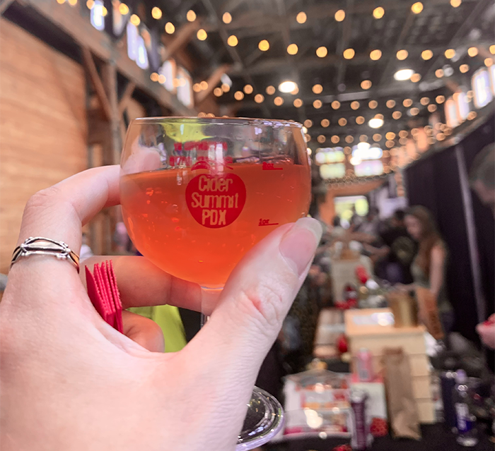 A hand holds a little Cider Summit tasting glass with Glow from Alpenfire Cider.