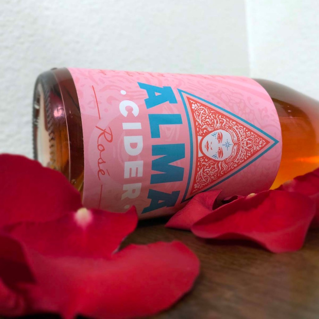 A bottle of Rosé from Alma Cider lays sideways surrounded by rose petals.