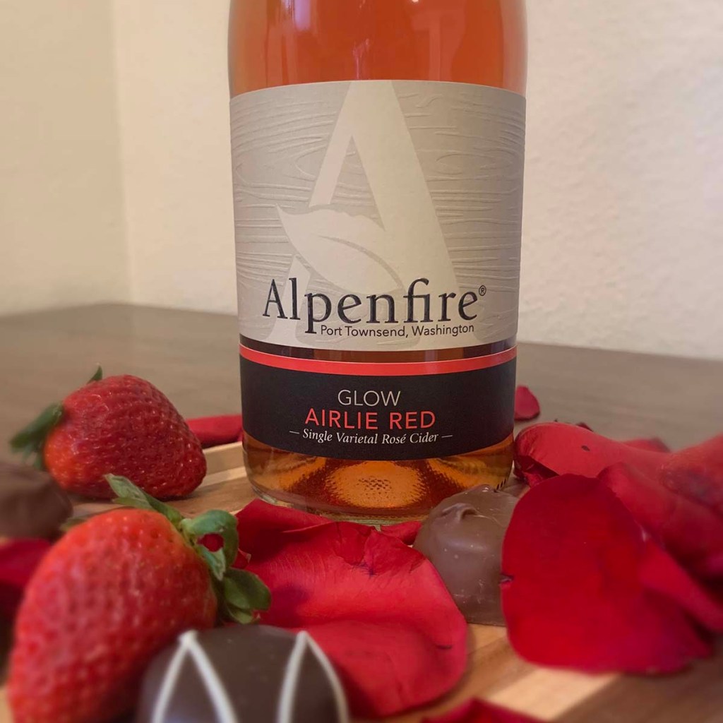 A bottle of Glow from Alpenfire Cider sits on a table surrounded by rose petals, chocolate and strawberries.