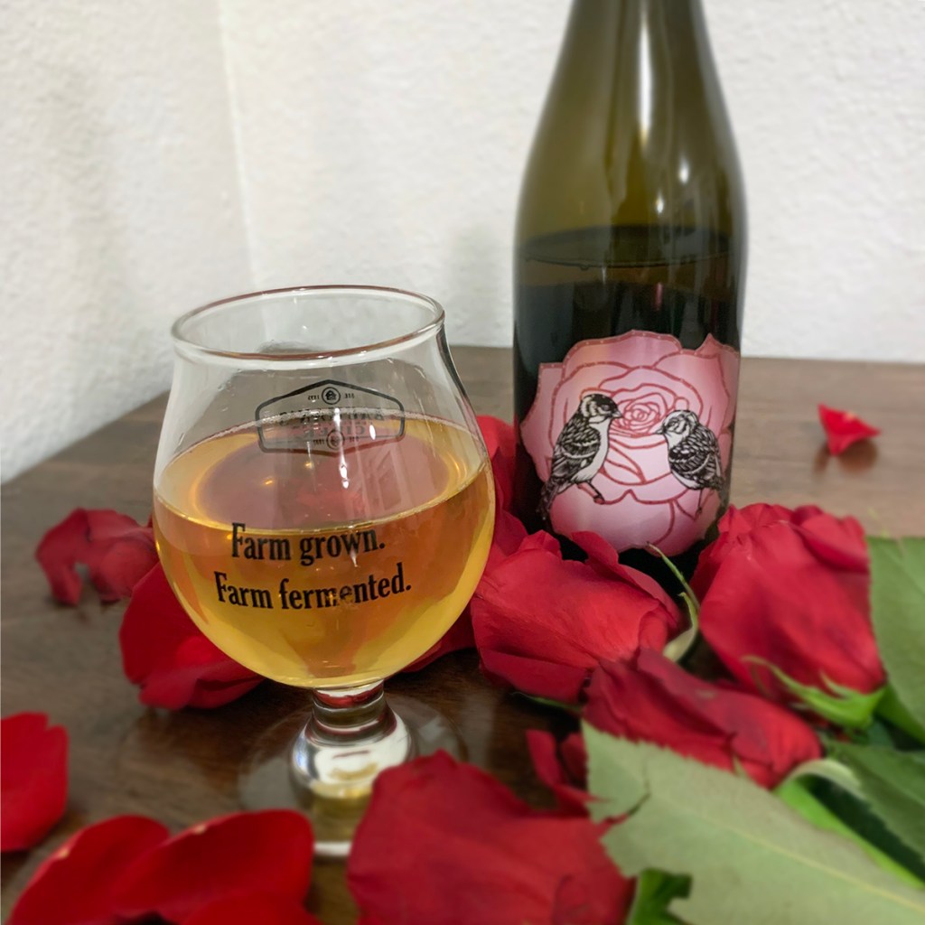 A glass of cider sits next to a bottle of 2018 Mountain Rose Single Varietal Cider by Art + Science Cider, with roses on a table.