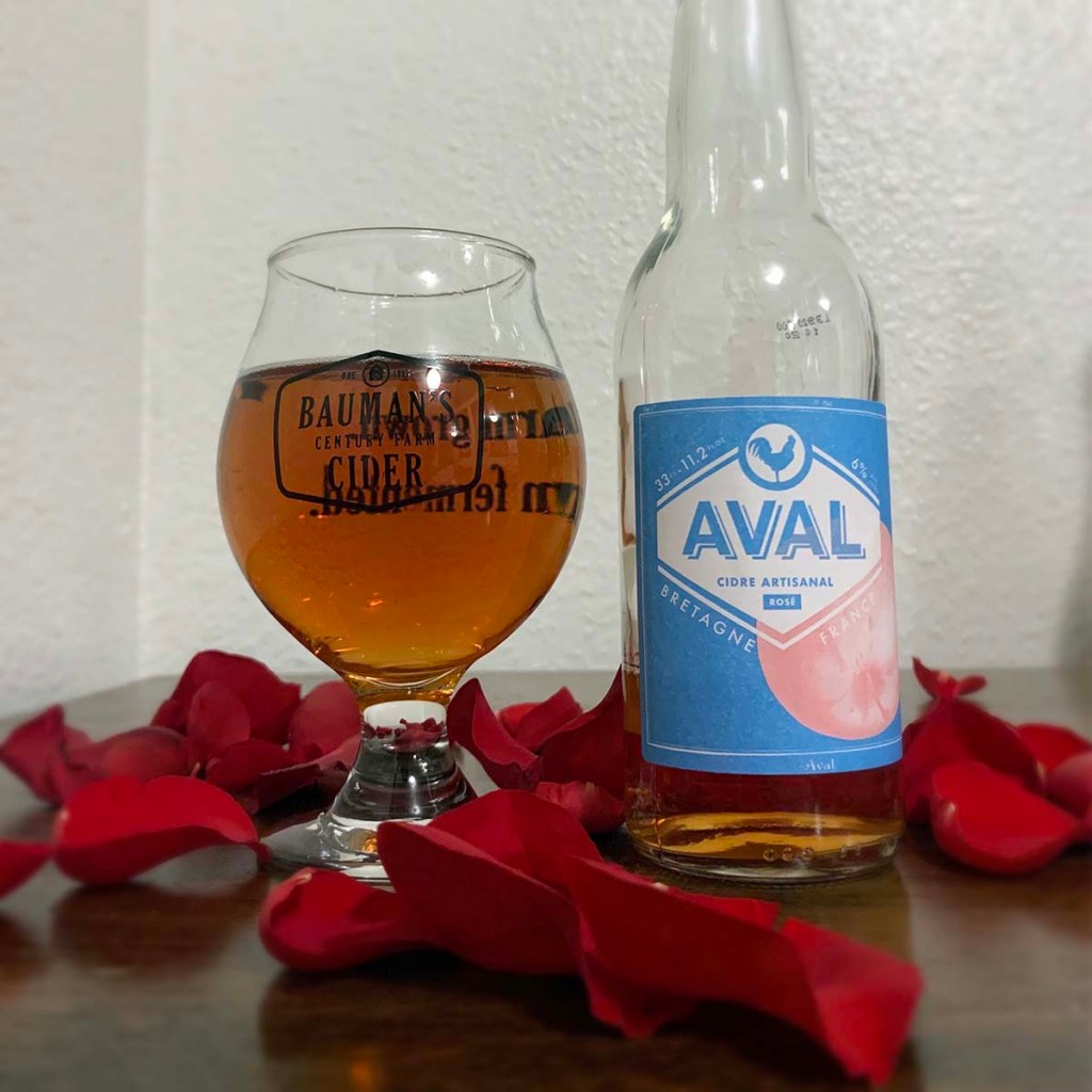 A glass of cider sits on a wood table next to a bottle of Rosé from Avalons Cider, surrounded by rose petals.