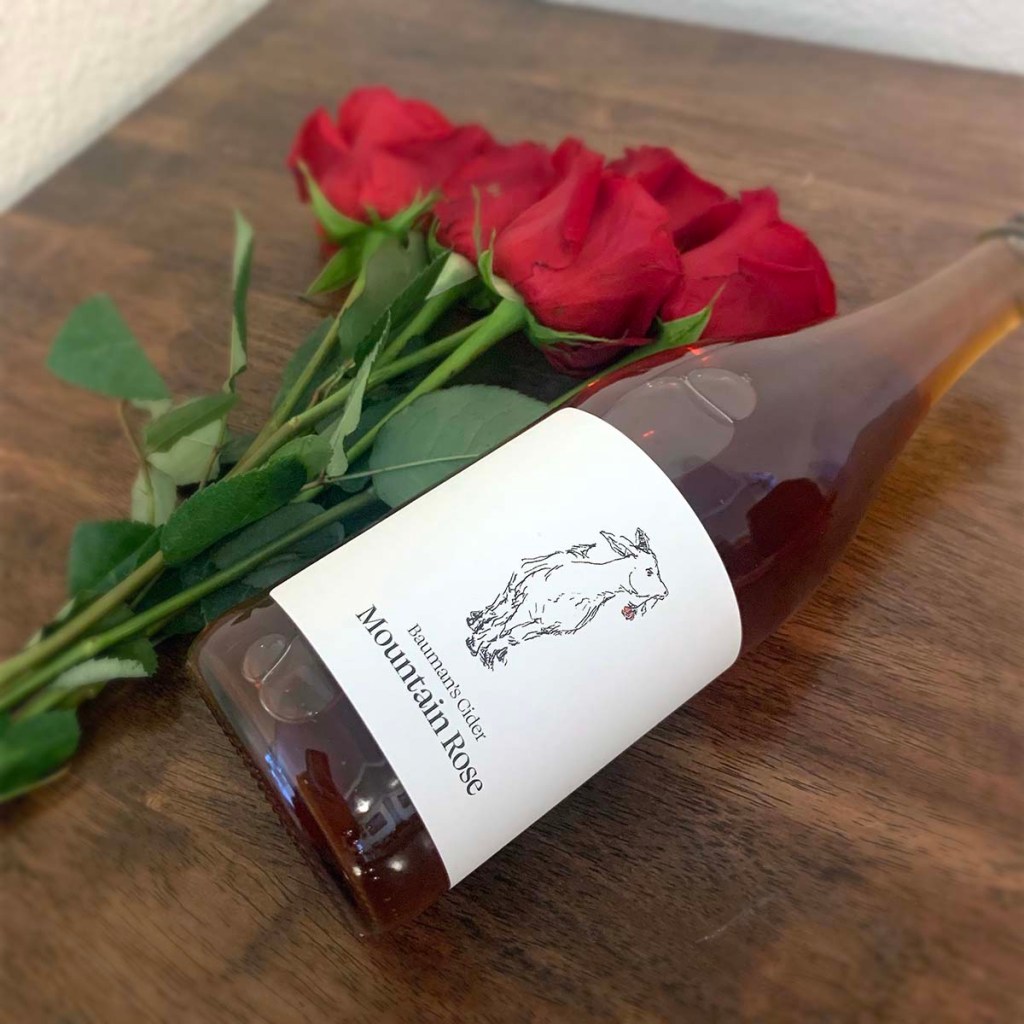 A bottle of Mountain Rose from Bauman's Cider lays sideways on a table next to some roses.
