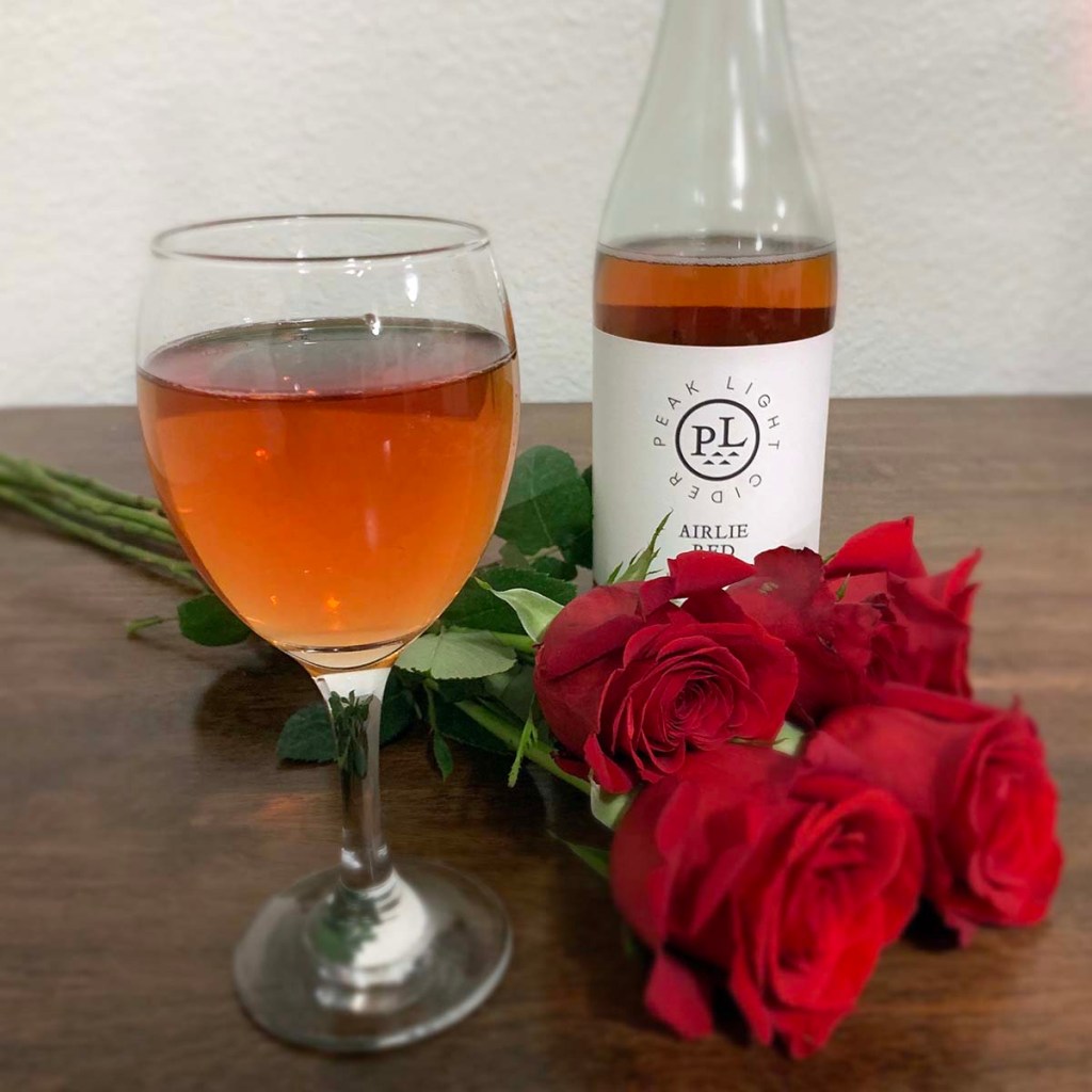 A glass of cider sits in front of a bottle of Airlie Red from Peak Light Cider and a bunch of roses.