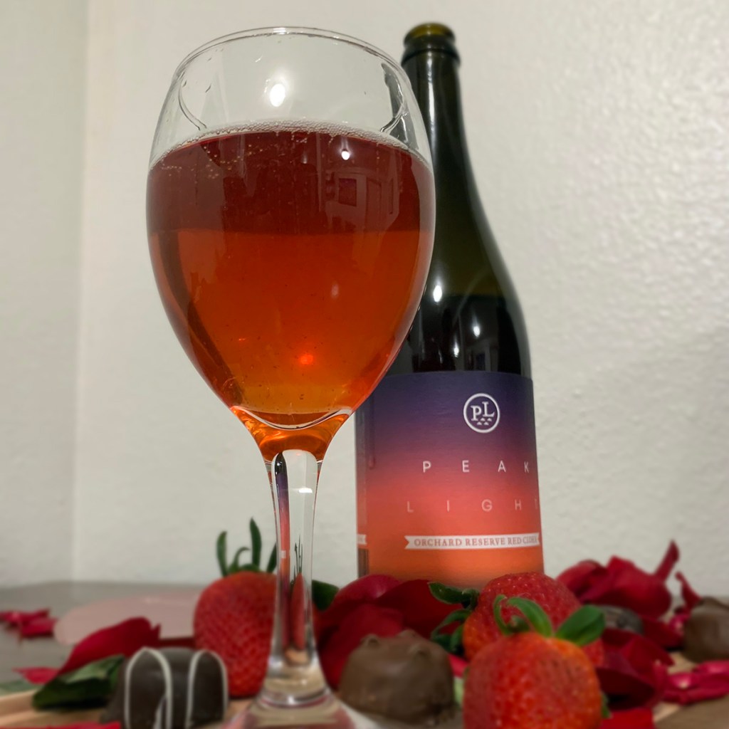 A glass of cider sits in front of a bottle of Orchard Reserve Red Cider from Peak Light Cider, surrounded by chocolates, rose petals and strawberries.
