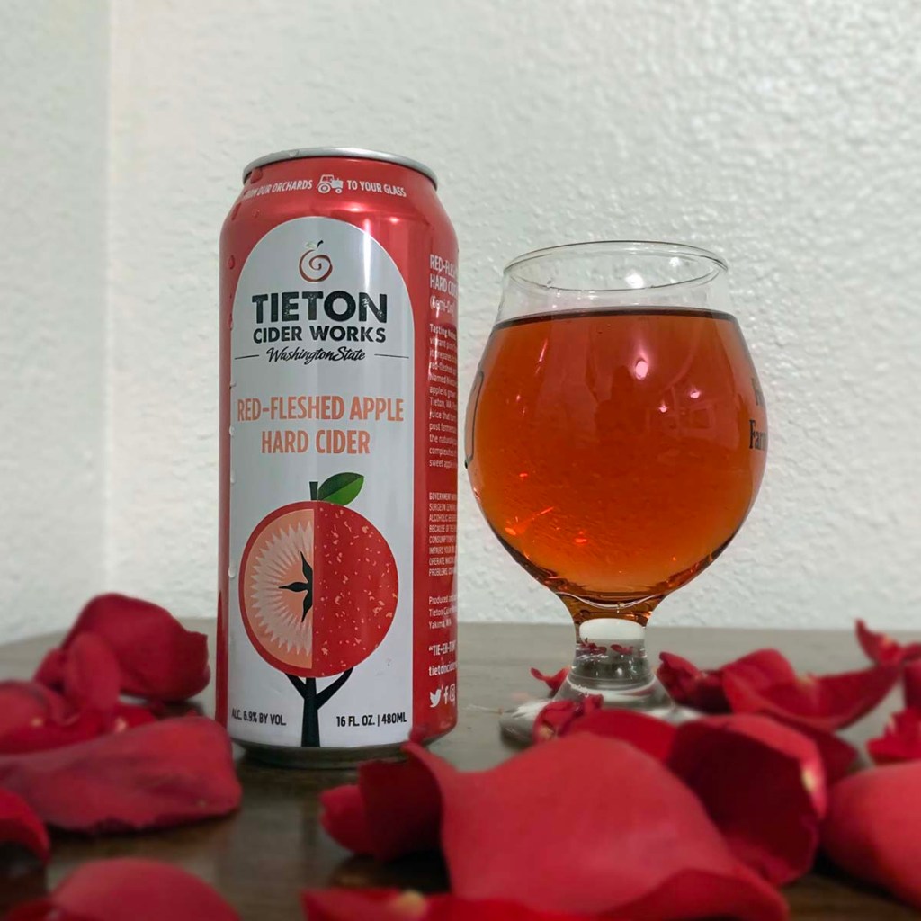 A glass of cider sits next to a can of Red-Fleshed Apple Hard Cider from Tieton Ciderworks, surrounded by rose petals.