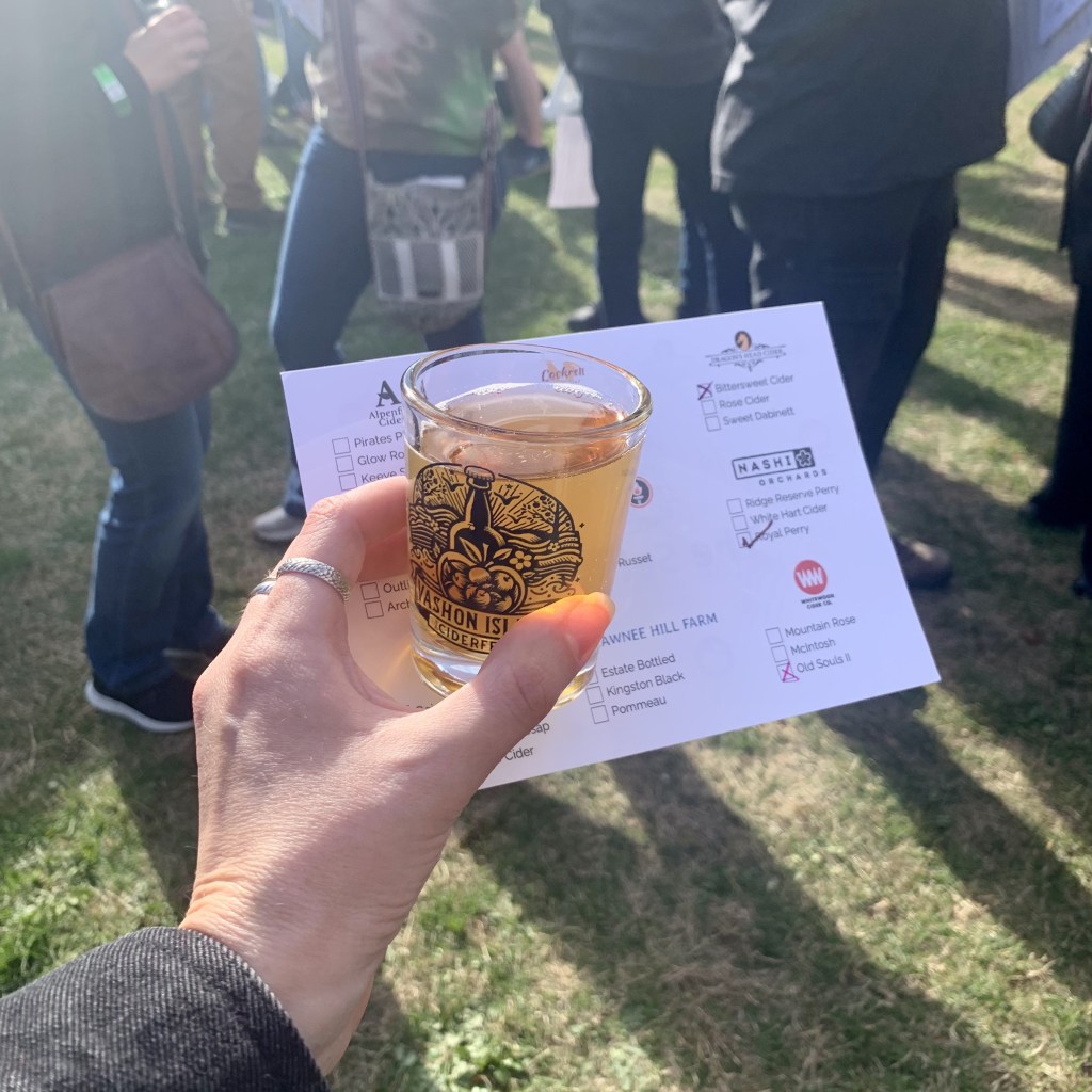 A hand holds a Vashon Island Cider Festival tasting glass filled with cider.