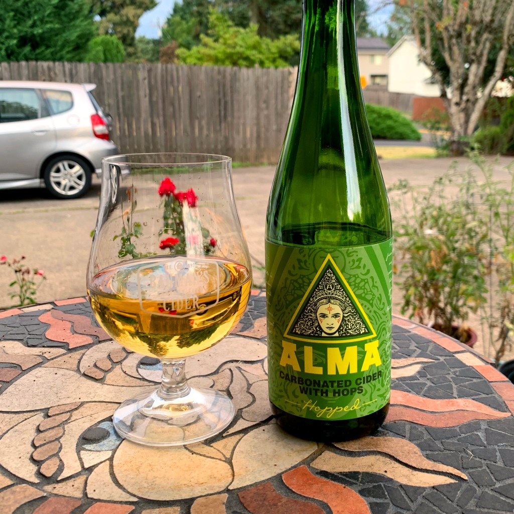 A glass of cider on a mosaic tiled table next to a bottle of Hopped Alma's Cider
