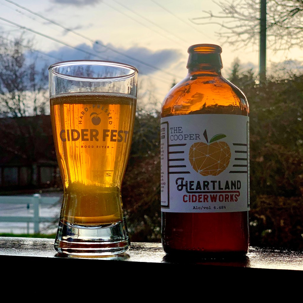 A glass of cider sits next to a bottle of The Cooper from Heartland Ciderworks