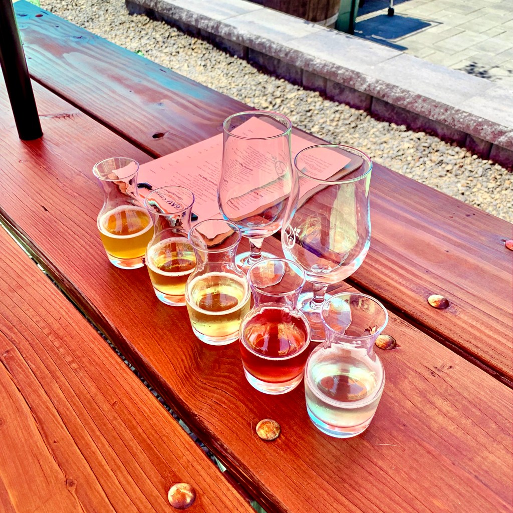 A cider flight from Raw Cider sits on a picnic table with a menu and two extra glasses